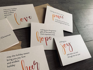 An angled, close-up photo of the 6 cards from the Meaning of Christmas collection lie on a gray wooden background with two stacks of kraft envelopes. The cards are printed on a cream pearlized paper. Each card has a large script word in an ombre of orange to burgundy. The rest of the sentiment is in black sans serif font.
