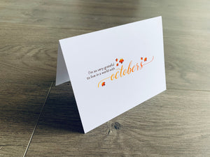 A white folded notecard is propped up on a wooden floor. The card says, "I'm so very grateful to live in a world with octobers" with small leaves around it. From the I Love Fall Collection by Stationare.