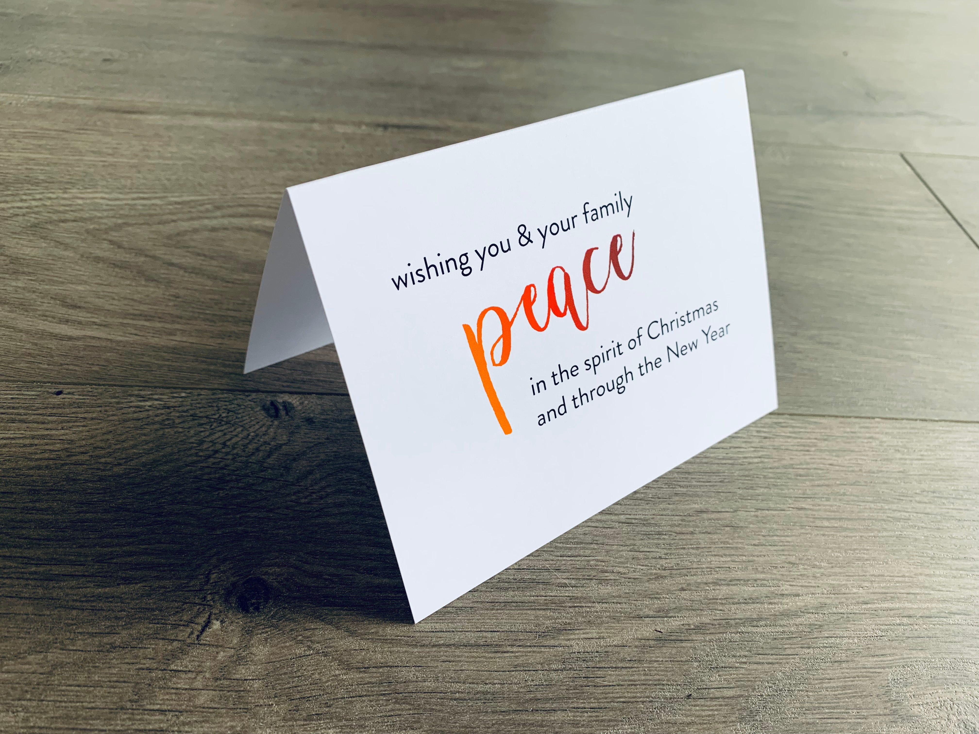 A white folded notecard is propped up on a wooden floor. The card says, "wishing you and your family peace in the spirit of Christmas and through the new year." From the Meaning of Christmas Collection by Stationare.