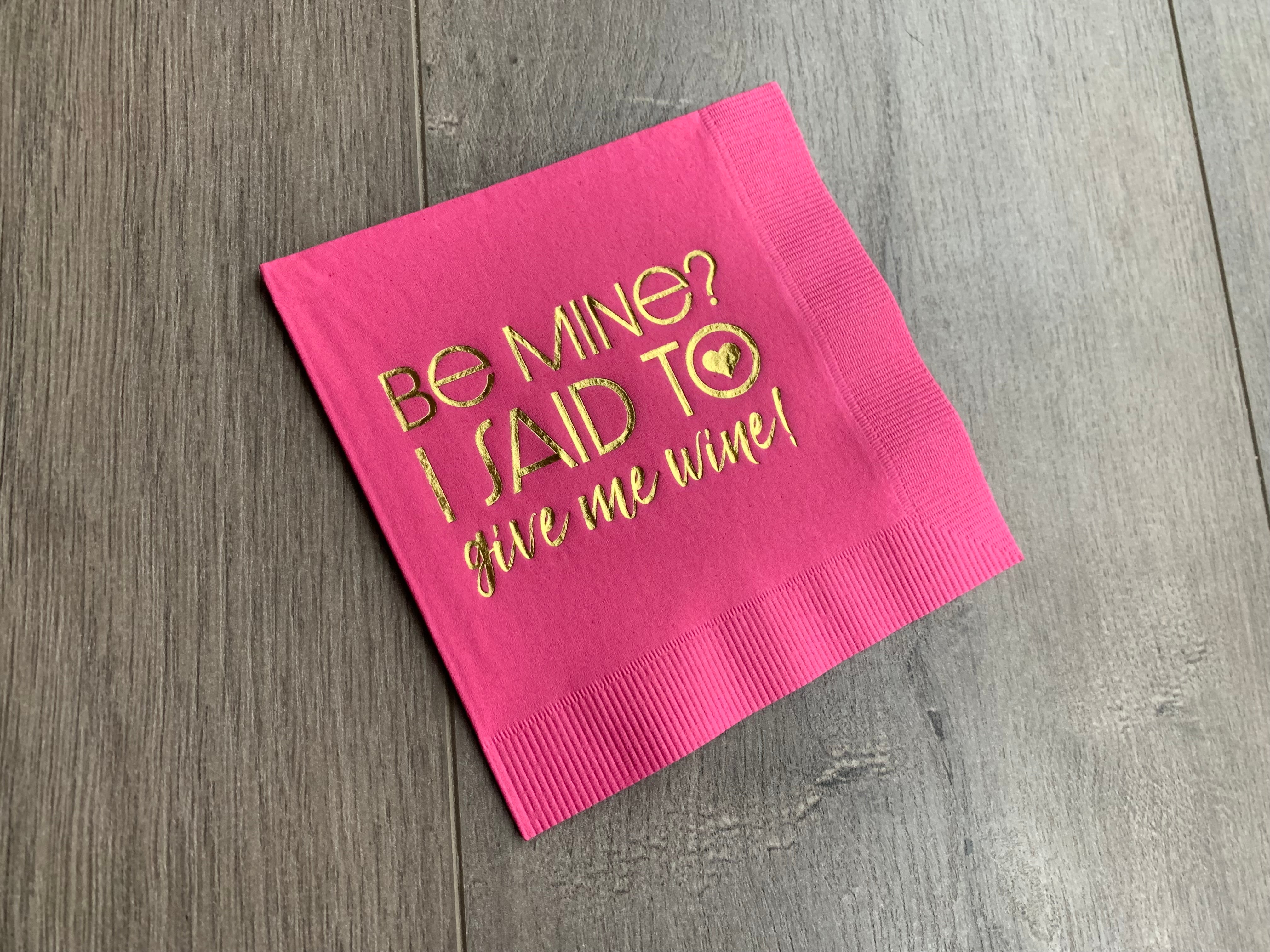 pink cocktail napkin on a wooden background. Metallic gold foil printing that reads Be Mine? I said to give me wine! Valentines's Day napkin by Stationare for girls night party, galentines, or a valentine party