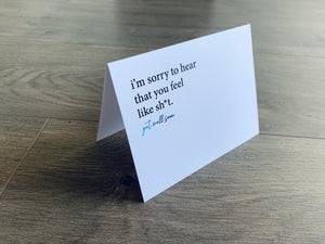 A white get well soon inspired notecard is propped up on a wooden floor. The card reads, "I'm sorry to hear that you feel like sh*t. Get well soon." From Stationare's joyful well wishes collection.
