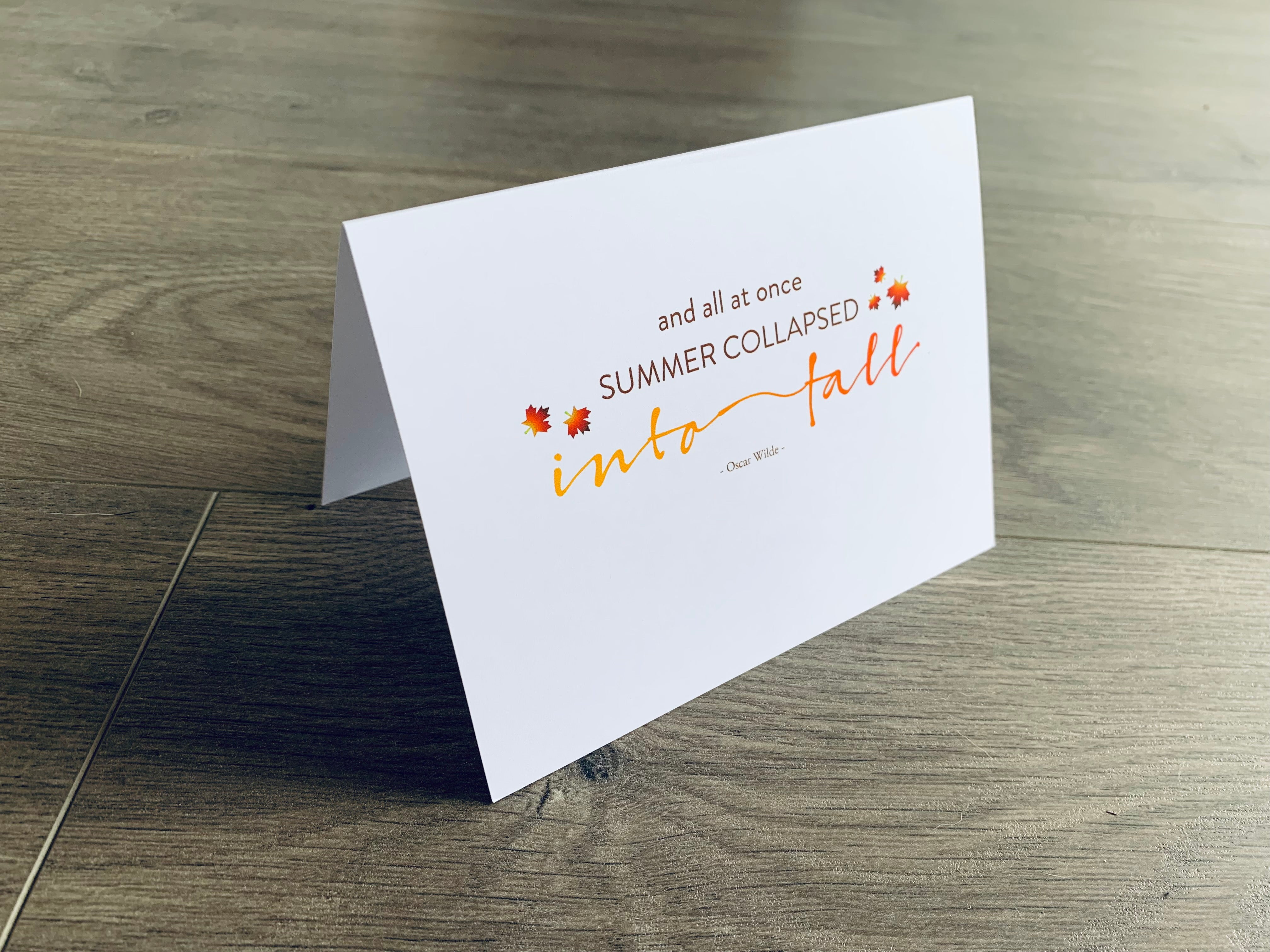 A white folded notecard is propped up on a wooden floor. The card says, "and all at once summer collapsed into fall" with small fall leaves around it. From the I Love Fall Collection by Stationare.
