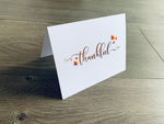 A white folded notecard is propped up on a wooden floor. The card says, "thankful" with small leaves around it. From the Giving Thanks Collection by Stationare.