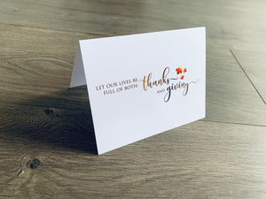 A white folded notecard is propped up on a wooden floor. The card says, "Let our lives be full of both - thanks and giving" with small leaves around it. From the Giving Thanks Collection by Stationare.