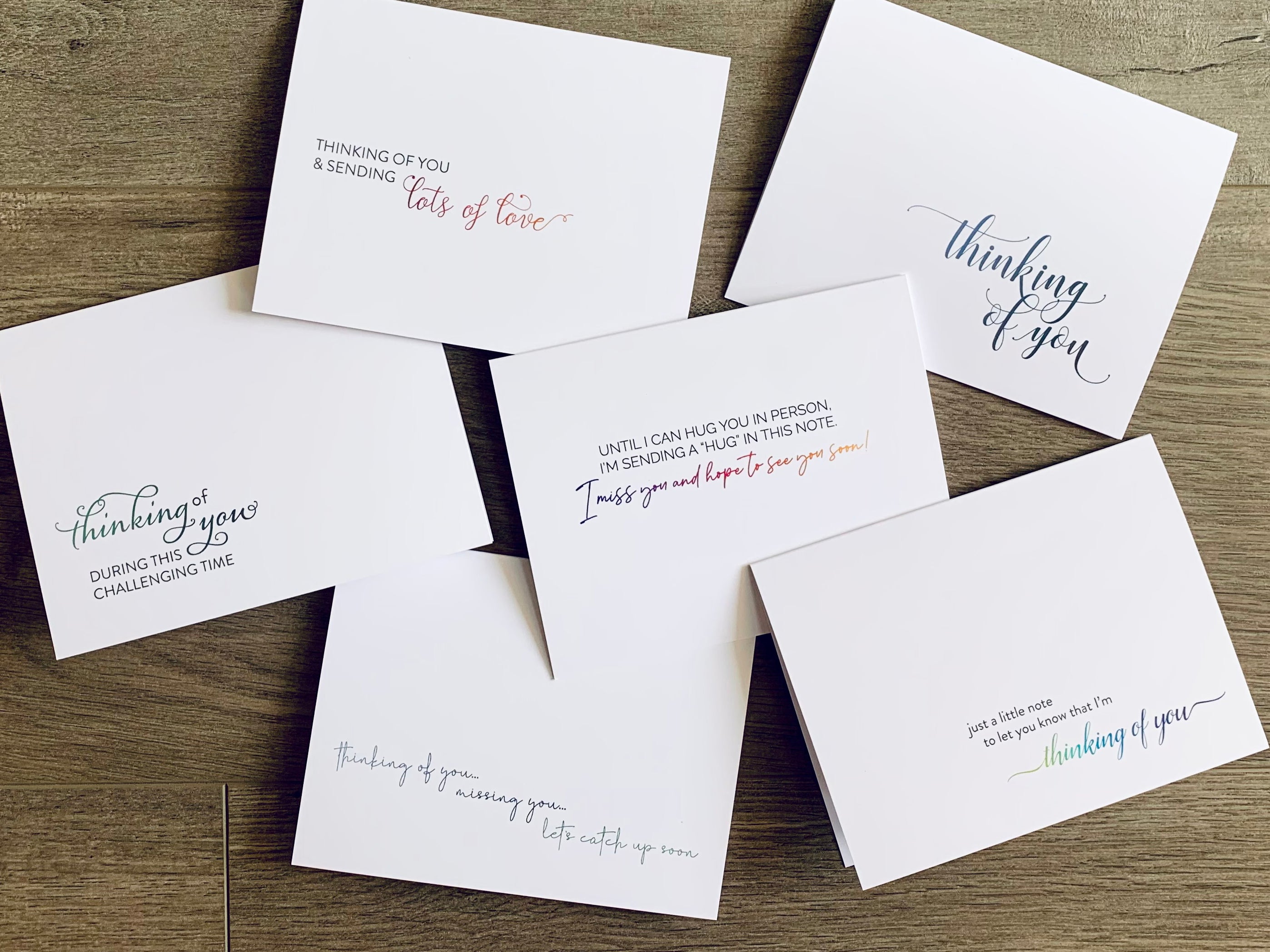 Six white notecards are overlapped and arranged on a wooden background. Each card has a mix of sans serif and script fonts with a thinking of you style sentiment. Thinking of You collection by Stationare.