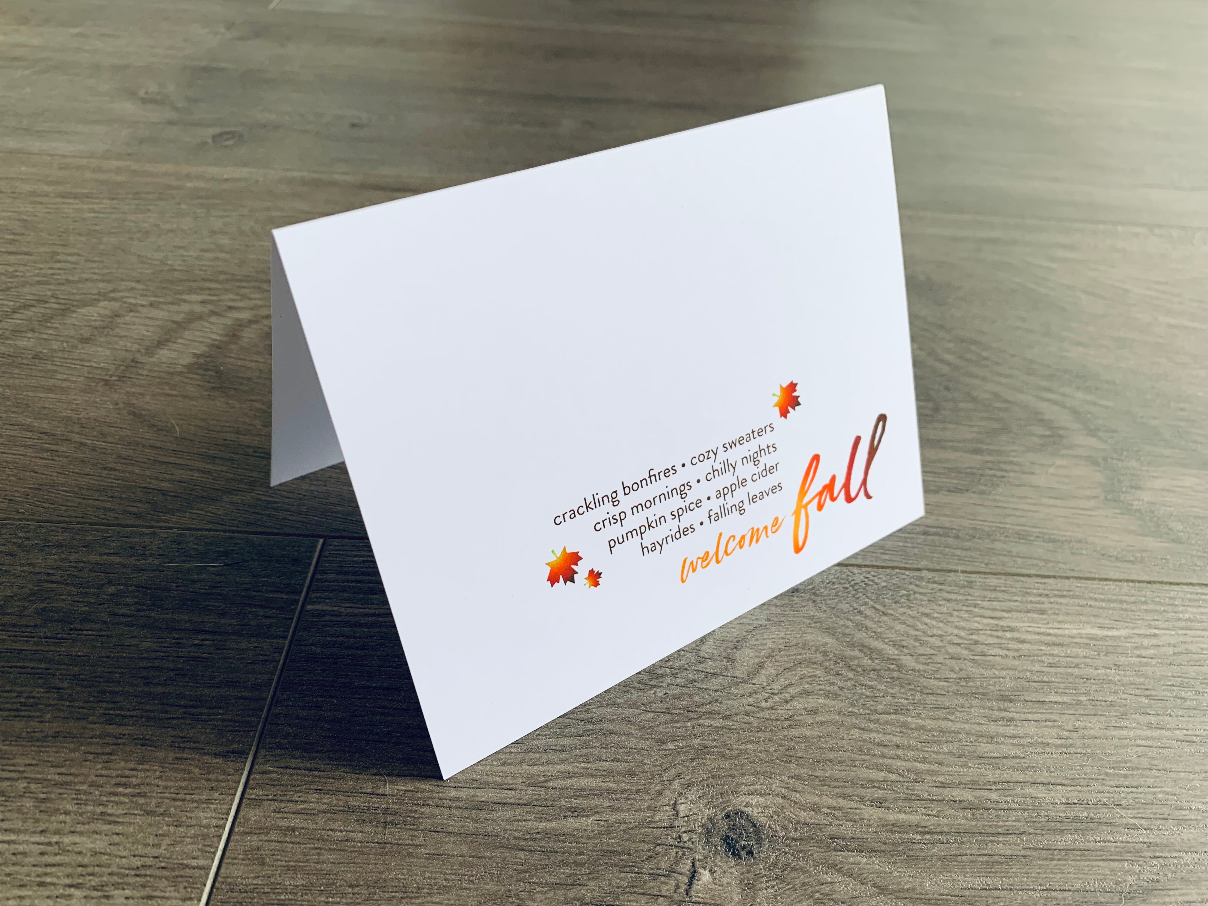 A white folded notecard is propped up on a wooden floor. The card says, "crackling bonfires. cozy sweaters. crisp mornings. chilly nights. pumpkin spice. apple cider. hayrides. fall leaves. welcome fall" with small fall-themed leaves around it. From the I Love Fall Collection by Stationare.