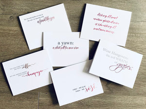 Six white notecards overlapped and lying on a wooden background. In a mix of script and serif fonts, there are quotes and sayings about wine and friendship in pink, gray and black ink. Wine Notecards by Stationare.