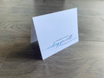A white, folded notecard is propped up on a gray wooden floor. Toward the bottom, it reads, "You've been in my thoughts... wishing you well and hoping for a speedy recovery." From Stationare's Get Well Soon collection.