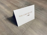 A white, folded notecard sits on a wooden floor. The card says, "You can do it. - coffee." Coffee Lovers collection by Stationare.