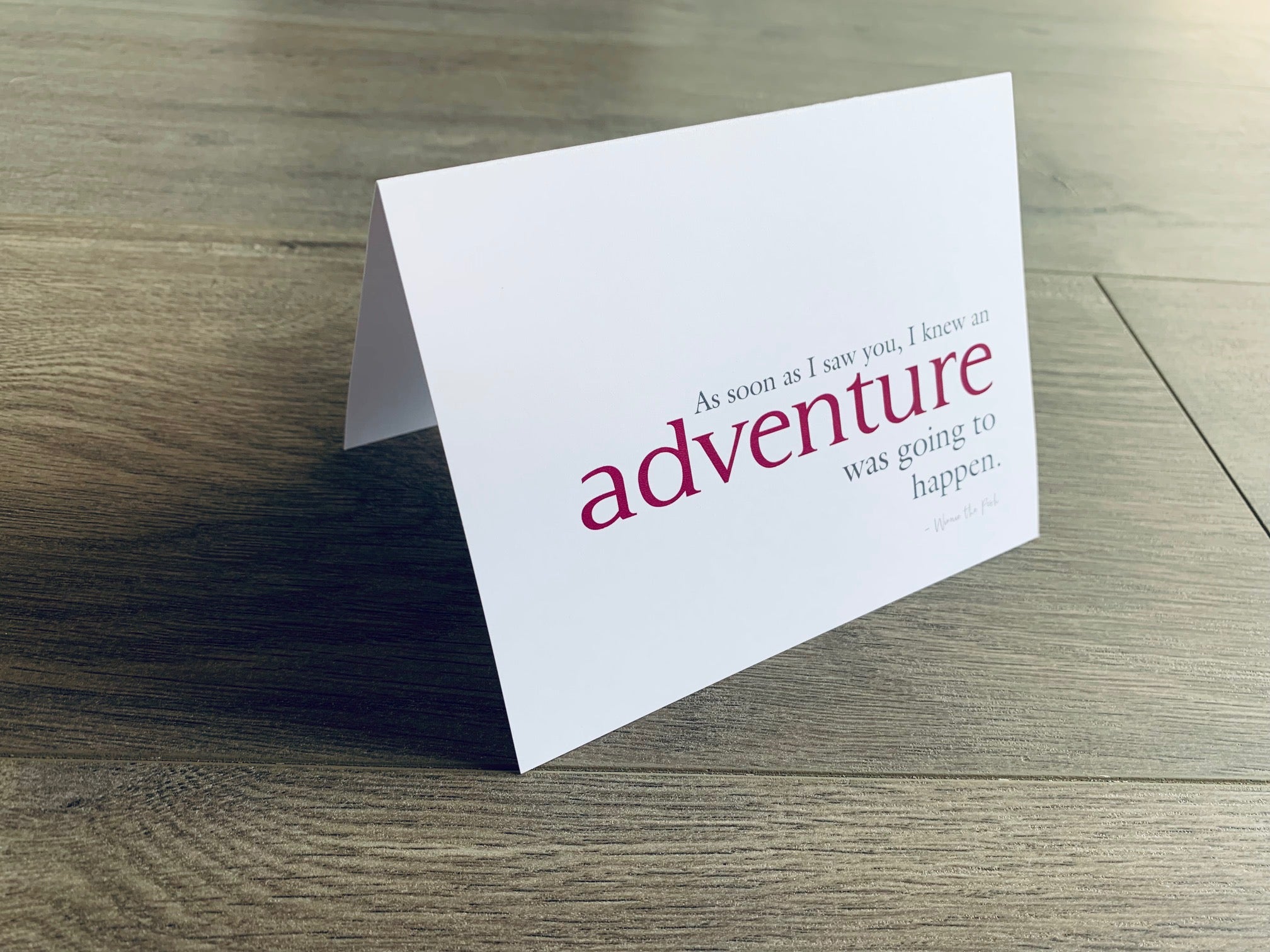 A white, folded notecard sits on a wooden floor. The card says, "As soon as I saw you, I knew an adventure was going to happen." The Friendship collection by Stationare.