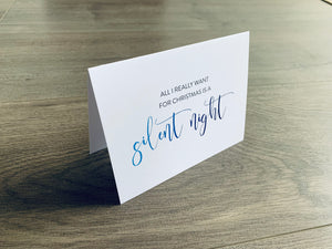 A white folded notecard is propped up on a wooden floor. On the front of the card, it says "All I really want for Christmas is a silent night." Christmas Chuckles collection by Stationare.