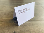 A white, folded notecard is propped up on a wooden background. In script font, it reads "All you need is love... and anything with chocolate." Card by Stationare.