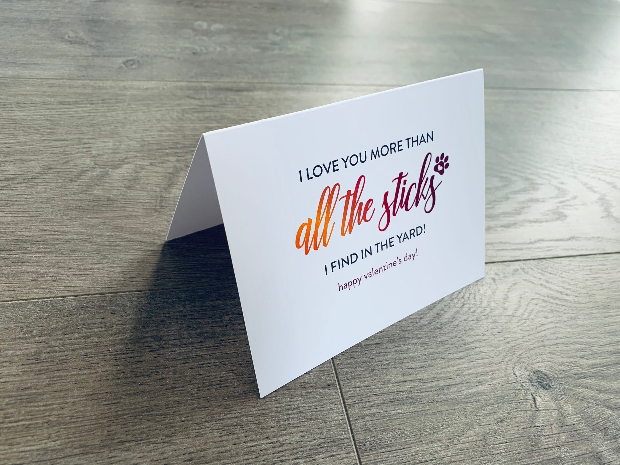 A folded white notecard is folded and propped up on a gray wooden background. The card says, "I love you more than all the sticks I find in the yard! Happy Valentine's Day!" Valentines from the Pup collection by Stationare.