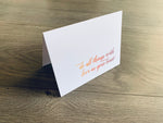 A folded white notecard is propped up on a wooden floor. The card reads, "do all things with love in your heart" in a script font. Love Collection by Stationare.