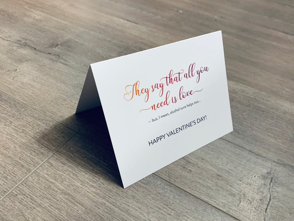 A folded white notecard is folded and propped up on a gray wooden background. The card says, "They say that all you need is love - but I mean, alcohol sure helps too... Happy Valentine's Day." Valentine Smiles collection by Stationare.