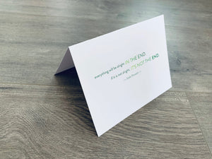 A white folded notecard sits on a gray wood floor. It reads, "Everything will be alright in the end. If it's not alright, it's not the end." Irish Inspirations collection by Stationare.