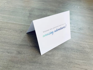 A white card is propped on a gray wood floor. The card reads, "Wishing you another  year of amazing adventures!" Birthday card by Stationare.