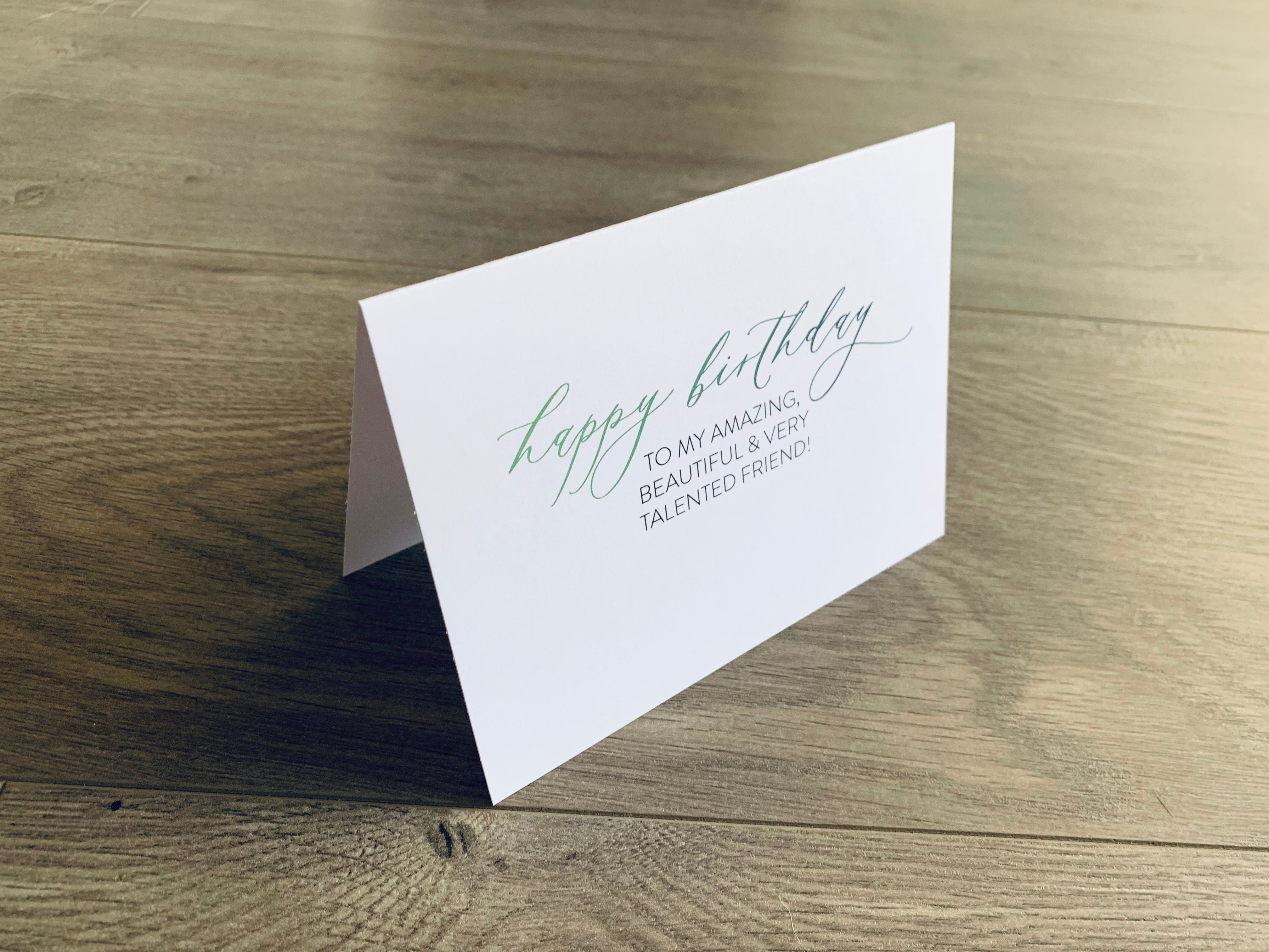 White folded notecard is propped up on a wooden floor. The card reads, "happy birthday to my amazing, beautiful and talented friend!" in a mix of script and serif fonts. From Stationare's Friendship Birthday collection.