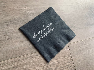 Angled black cocktail napkin lying on a wooden floor. In white script writing, it reads, "cheers, cheese & charcuterie" by Stationare