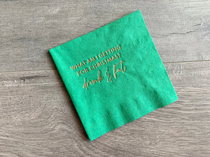 Angled green Christmas napkin on a gray wooden background. In champagne foil it reads, " What am I getting for Christmas? drunk & fat." By Stationare