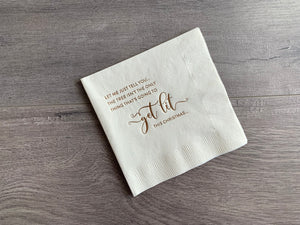 Angled, off-white cocktail napkin lies on a gray wood background. In champagne foil it reads, "Let me just tell you, the tree isn't the only thing that's going to get lit this Christmas." 