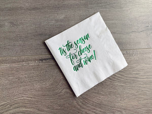An angled white napkin rests on a gray wooden background. In green metallic foil, it reads, "tis the season for cheese and wine" in a script font. By Stationare