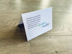 A white card is propped on a gray wood floor. The card reads, "Do we really get smarter as we get older? Or have we just worked through our list of stupid ideas? Either way, here's to another year of adventures and laughter!" Birthday Smiles collection by Stationare