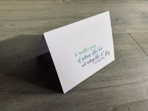 A white, folded birthday card is propped up on a gray wooden floor. The card reads, "to another year of getting older but not acting like it! Cheers!" in script font.  Birthday card by stationare, birthday cheers