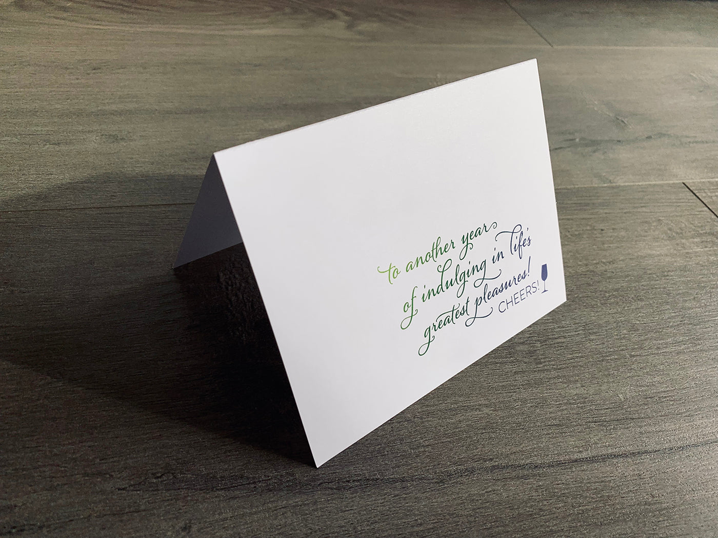 White folded notecard is propped up on a gray wooden floor. The card reads, "to another year of indulging in life's greatest pleasures! cheers!" From Stationare's Birthday Cheers collection.