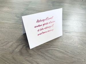 A white notecard is folded and propped up on a wooden floor. The card says, "Asking if I want another glass of wine is like asking if I want more money." A wine-themed notecard by Stationare