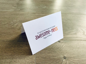 A white card is propped on a gray wood floor. The card reads, "To another year of sheer awesome-ness. Happy Birthday!" Birthday Inspirations by Stationare