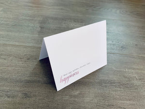 A white card is propped on a gray wood floor. The card reads, "flour. eggs. sprinkles. chocolate. sugar. happiness." Bakers Collection by Stationare.