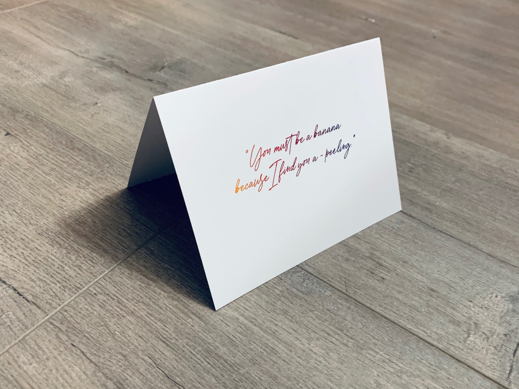 A folded white notecard is propped up on a wooden floor. The card reads, "You must be a banana because I find you a-peeling." Stationare's Pick Up Lines collection.