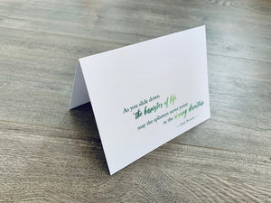 A white folded notecard is propped up on a wooden floor. The card has the Irish proverb, "As you slide down the banister of life, may the splinters never point in the wrong direction." From the Irish Laughs Collection by Stationare.