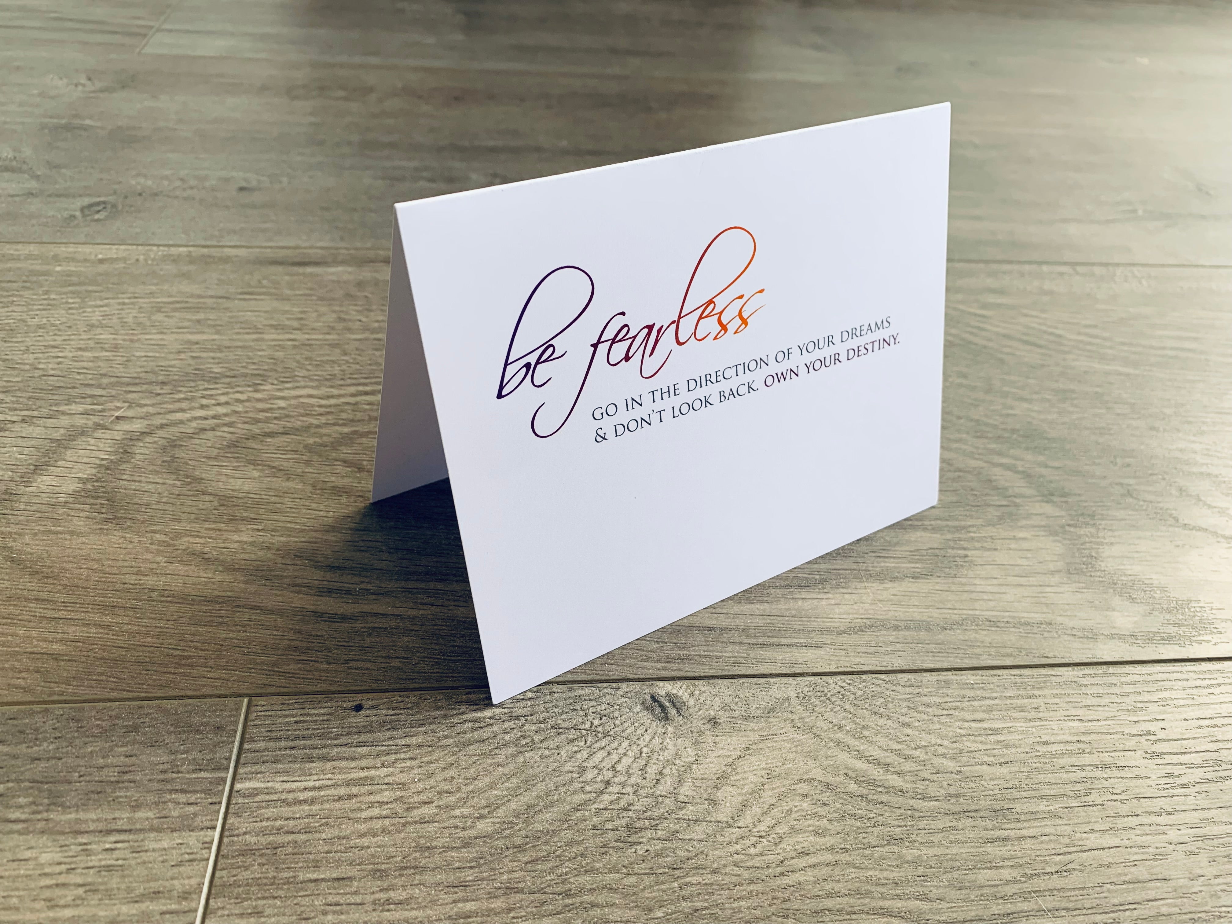 White folded notecard propped up on a wooden floor. The card reads "be fearless" in script font that fades from purple to orange. Below it is capitalized serif lettering that reads, "Go in the direction of your dreams and don't look back. Own your destiny." The card is from Stationare's Inspire collection.