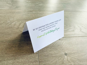 A white card is propped on a gray wood floor. The card reads, "Be so distracted living your life that you never notice that you're getting older. Happiest of birthdays to you." Birthday Inspirations by Stationare.