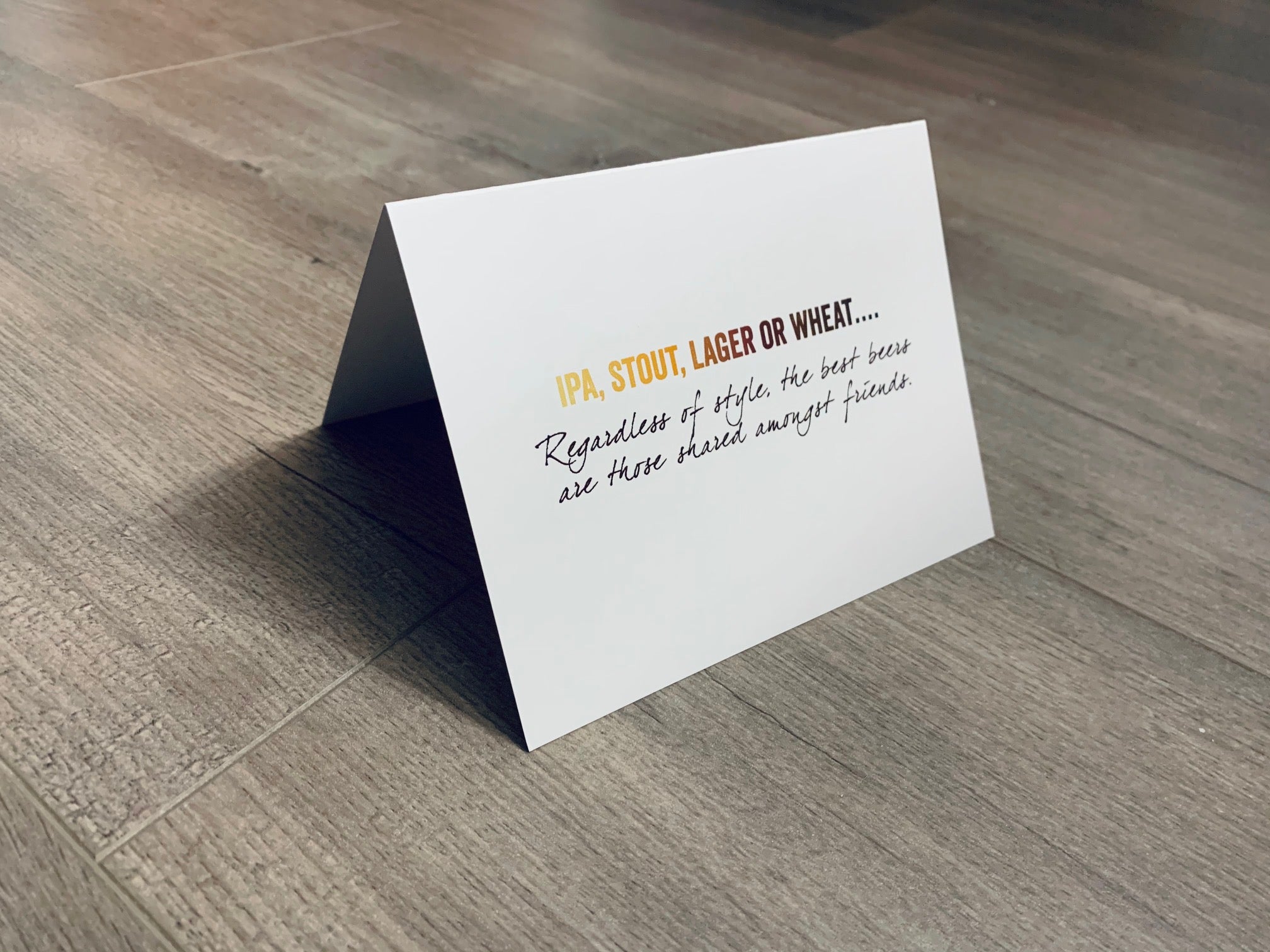 A white card is propped on a gray wood floor. The card reads, "IPA, stout, lager or wheat... Regardless of style, the best beers are those shared amongst friends." Beer Lovers Collection by Stationare.