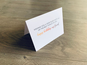 White folded notecard is propped up on a wooden floor. The card reads, "Wishing you a year filled with all of the best things in life! Happy birthday, my friend!" in a mix of script and serif fonts. From Stationare's Friendship Birthday collection.