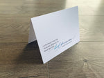 A white folded notecard stands on a gray wood floor. The card reads, "Let's make this the year we resolve to make better bad decisions. Happy New Year!"