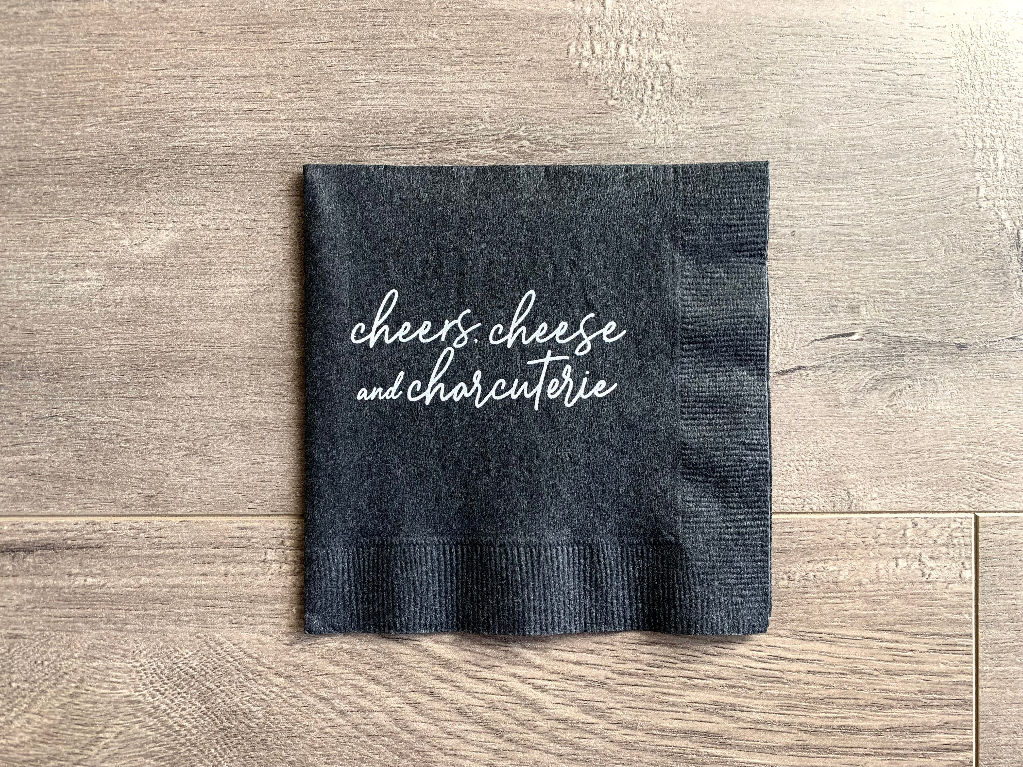 Black cocktail napkin lies on a wooden background. In matte ink, script writing reads, "cheers, cheese & charcuterie" by Stationare