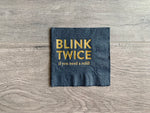 Black cocktail napkin with gold foil block printing that reads, "Blink Twice if you need a refill." It rests on a gray wooden background. By Stationare.