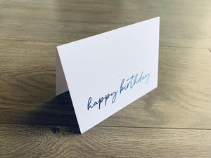 A single white notecard is propped up on a wooden floor. The card reads, "happy birthday" in a handwritten blue ombre font. Happy Birthday Collection by Stationare.