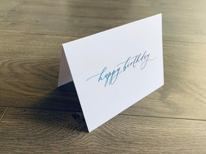 A single white notecard is propped up on a wooden floor. The card reads, "happy birthday" in a fancy script font in blue ombre. Happy Birthday Collection by Stationare.