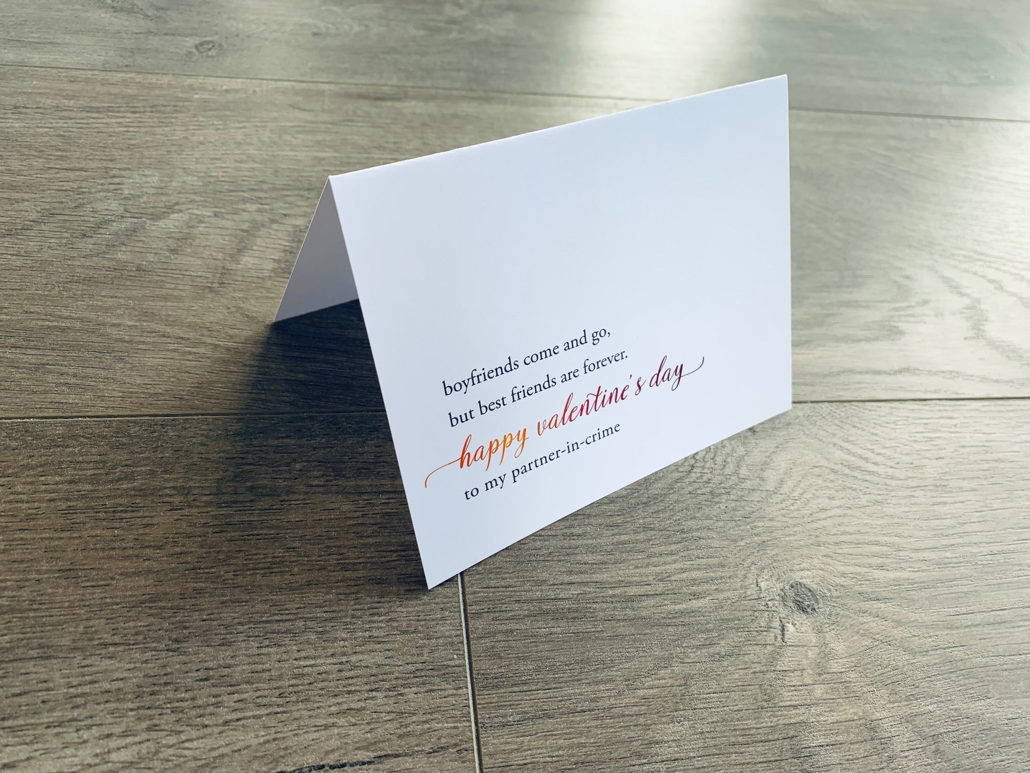A folded white notecard is folded and propped up on a gray wooden background. The card says, "Boyfriends come and go, but best friends are forever. Happy Valentine's Day to my partner-in-crime." VDay BFFs collection by Stationare.