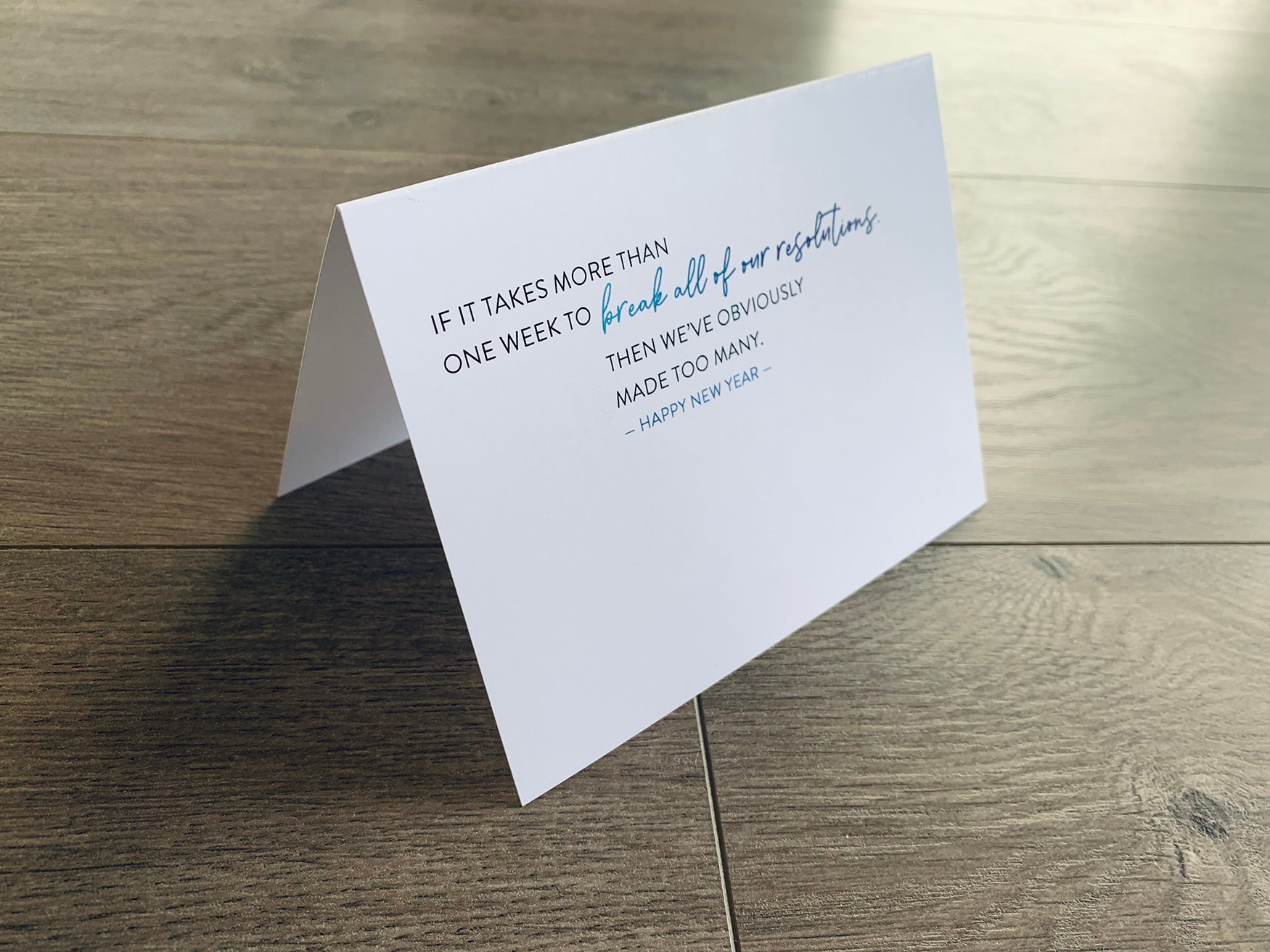 A white folded notecard stands on a gray wood floor. The card says, "If it takes more than one week to break all of our resolutions, then we've obviously made too many."