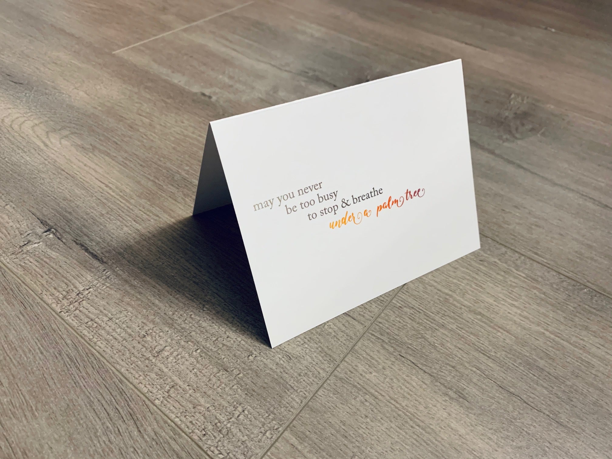 A white card is propped on a gray wood floor. The card reads, "May you never be too busy to stop and breathe under a palm tree." Beach Life Collection by Stationare.