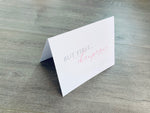 A white folded notecard is propped up on a gray wood floor. On the front of the card, it says, "But first... champagne." Champagne Lovers collection by Stationare.