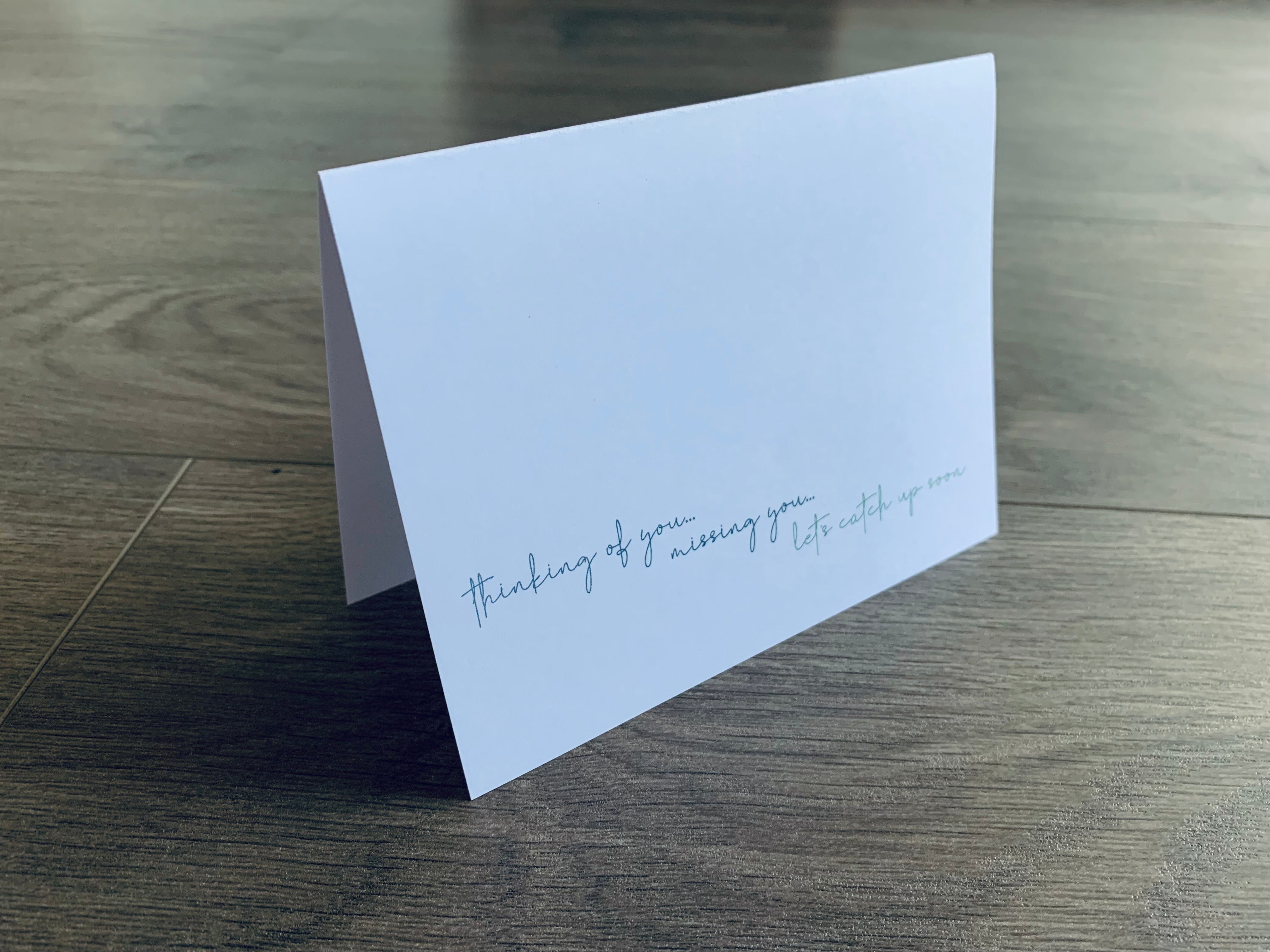 A white notecard is propped up on a gray wooden floor. The card reads, "Thinking of you. Missing you. Let's catch up soon." Stationare's Thinking of You collection.