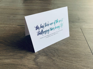 A folded white notecard is propped up on a wooden floor. The card reads, "this has been one of the most challenging times in my life. Thank you for helping me through it. I'm not sure what I would have done without you." Stationare's Surviving Hard Times collection.