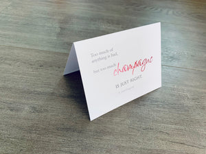 A white notecard is on a gray wooden floor. The card says,"Too much of anything is bad, but too much champagne is just right." Champagne Lovers collection by Stationare.